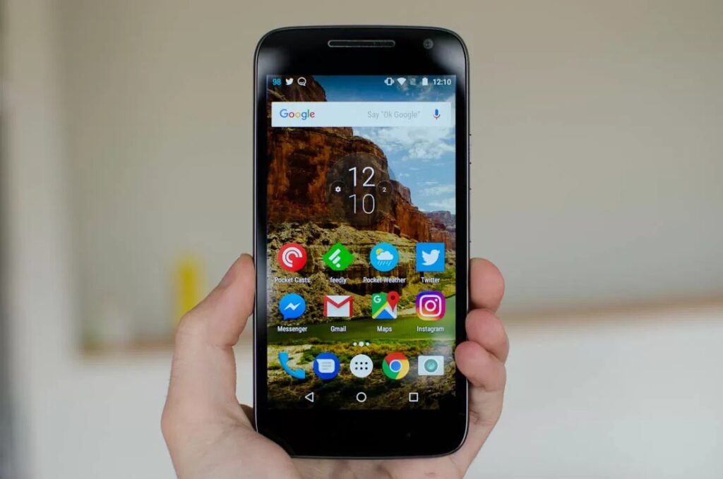Hand holding Motorola Moto G4 Play with Android home screen displaying Google search bar, clock, and popular app icons.