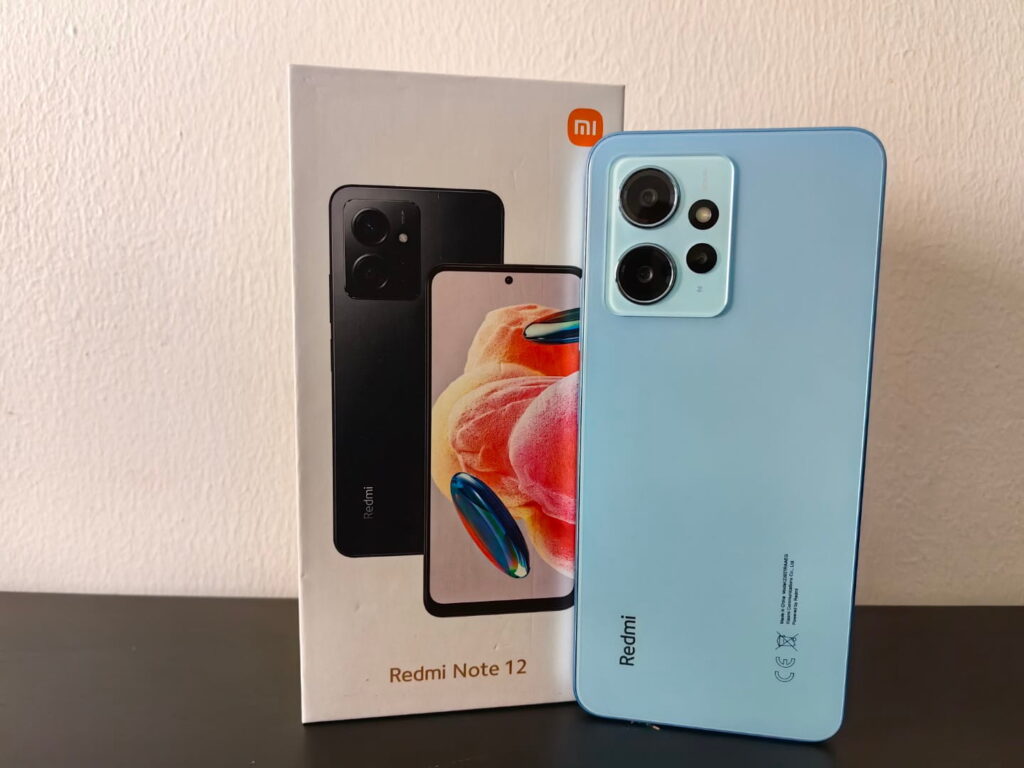 Redmi Note 12 4G in ice blue color leaning against its retail box on a dark surface, showing the rear triple-camera module, with the Redmi Note 12 box displaying the black color variant and Xiaomi Mi logo.