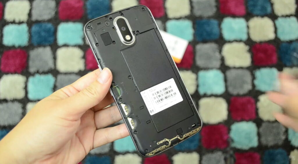 Hand holding a disassembled Motorola Moto G4 smartphone with the back cover removed, exposing the internal frame, camera module, fingerprint sensor, and barcode label, against a colorful checkered rug background.